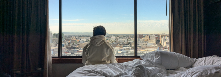 A view of someone sitting on an unmade bed, looking out the window, back to the camera, with a view of buildings outside the window